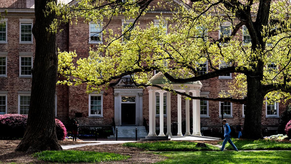 Wide-angle shot of the Old Well on the campus of UNC-Chapel Hill with a student walking by it. 南楼在背景中.