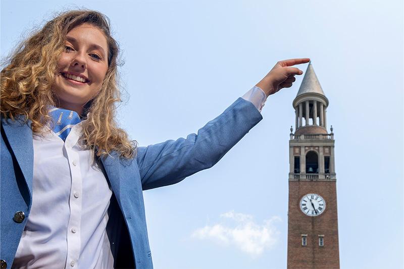 A student posing for a photo with the Bell Tower in the background. She's placing her finger to make it seem it's on top of the Bell Tower.